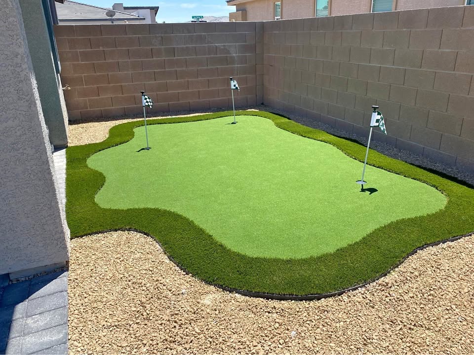 A small backyard putting green with three golf flags, surrounded by artificial grass and a border of light-colored gravel, enclosed by beige brick walls.