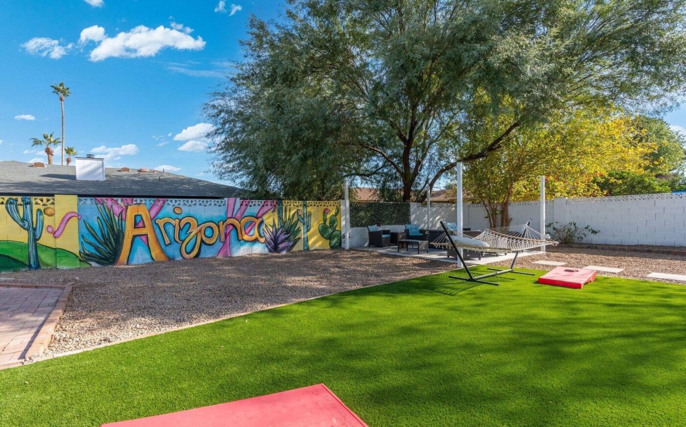 A backyard with artificial grass, a hammock, red cornhole boards, outdoor seating, and a mural on the back wall that reads "Arizona" with desert-themed artwork and cacti, under a sunny blue sky.