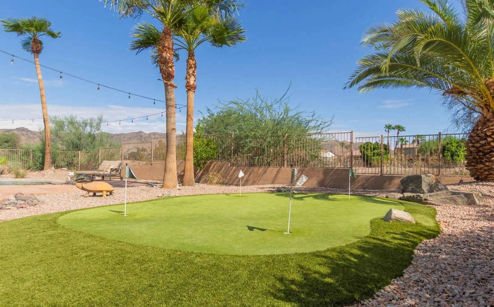 A small outdoor putting green with three golf flags is surrounded by palm trees, desert landscaping, rocks, and a pool. Mountains are visible in the background under a clear blue sky.