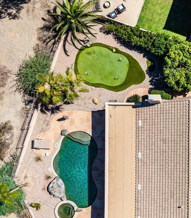 Aerial view of a backyard with a swimming pool, hot tub, putting green, palm trees, lounge chairs, and a tiled patio next to a house with a tan roof.