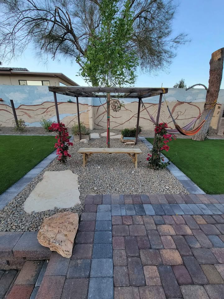 A backyard garden by Avondale Turf and Landscape features a wooden bench under a flower-covered pergola. A stone path winds through green grass, decorative rocks, a colorful hammock, and a mural brightens the wall in the background.