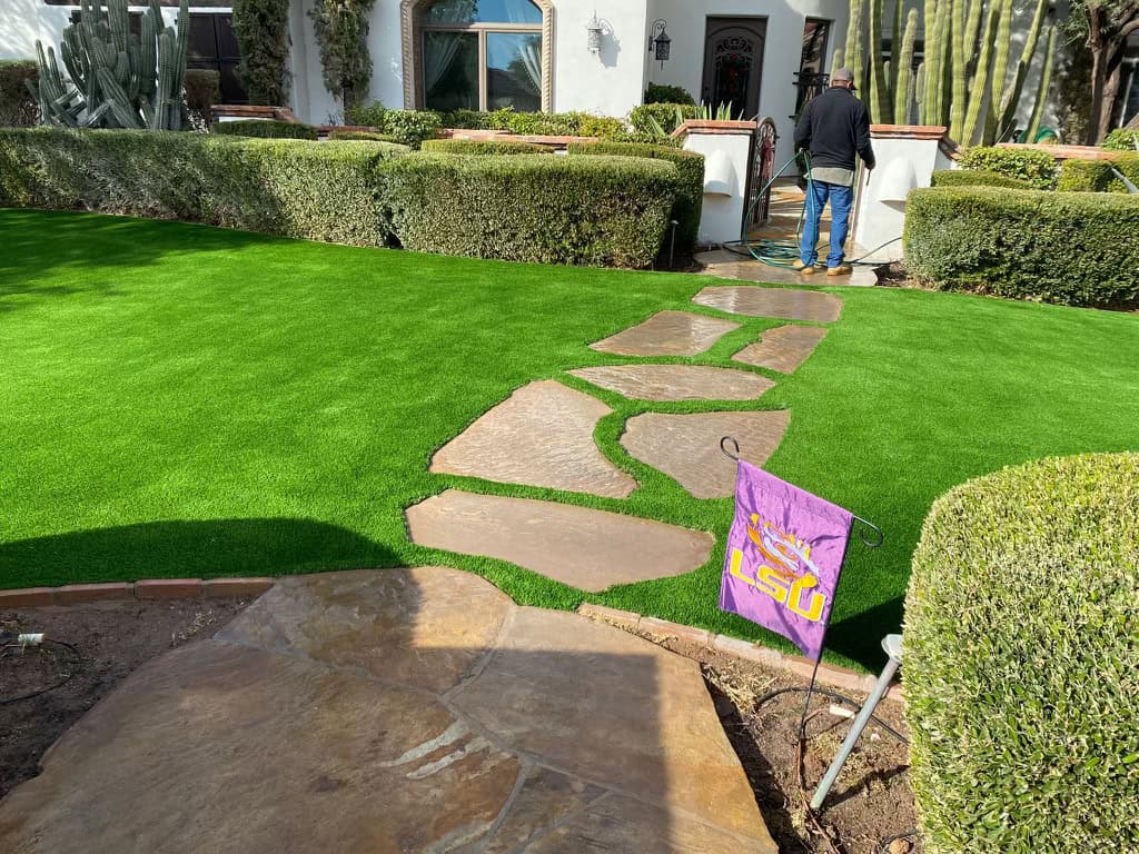 A person waters plants in front of a house with trimmed bushes, a vibrant green lawn by Avondale Turf and Landscape, a stone pathway, and a small purple LSU flag near the walkway.