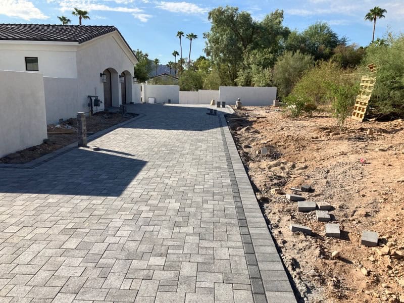 A newly installed gray stone paver driveway by Avondale Turf and Landscape sits beside a white house, with loose pavers and dirt along the right side, and trees and plants in the background under a sunny sky.