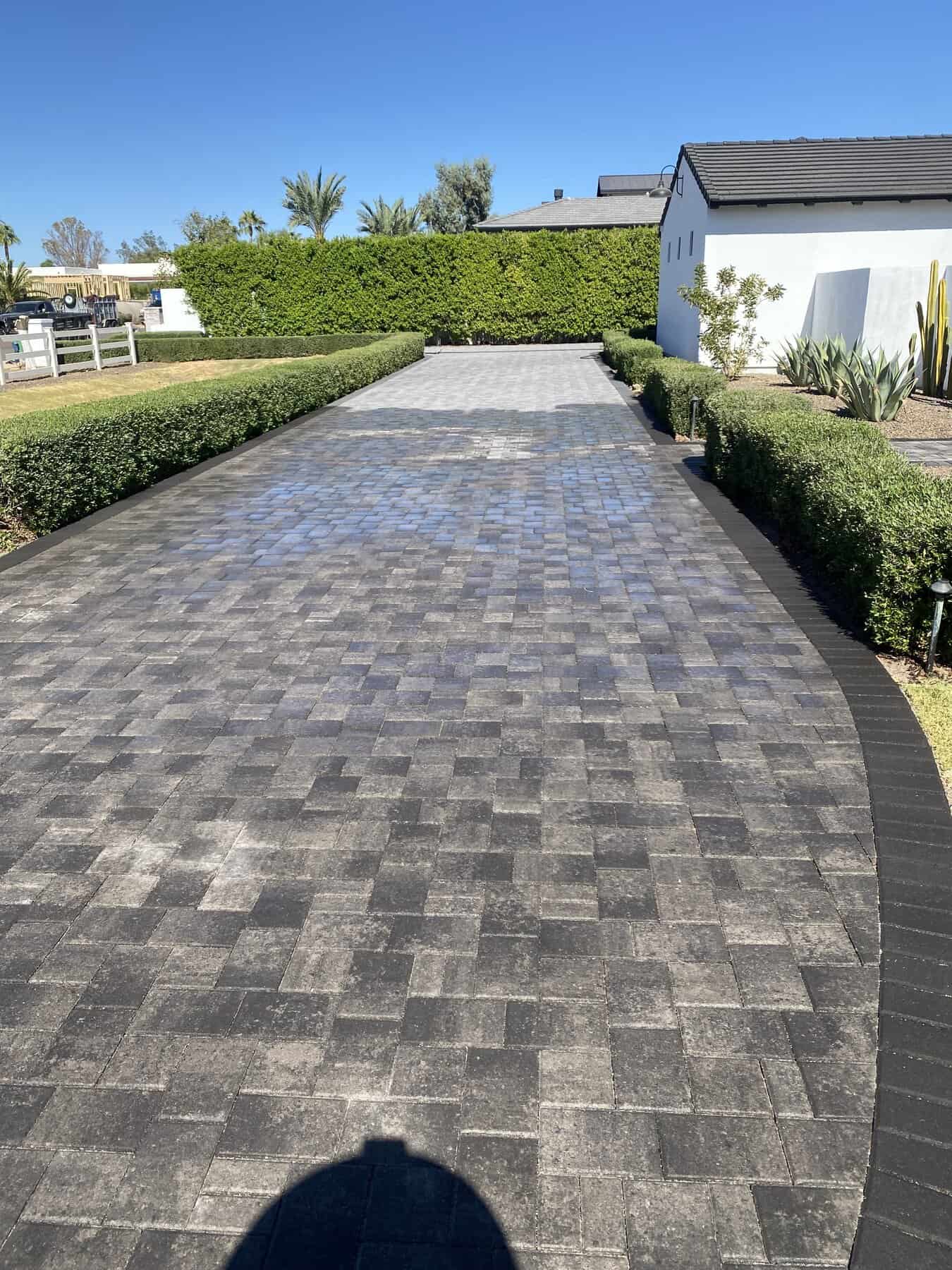 A paved driveway with a gradient from dark to light gray bricks, lined with green bushes and plants by Avondale Turf and Landscape, leads to a white house with a dark roof on a sunny day.