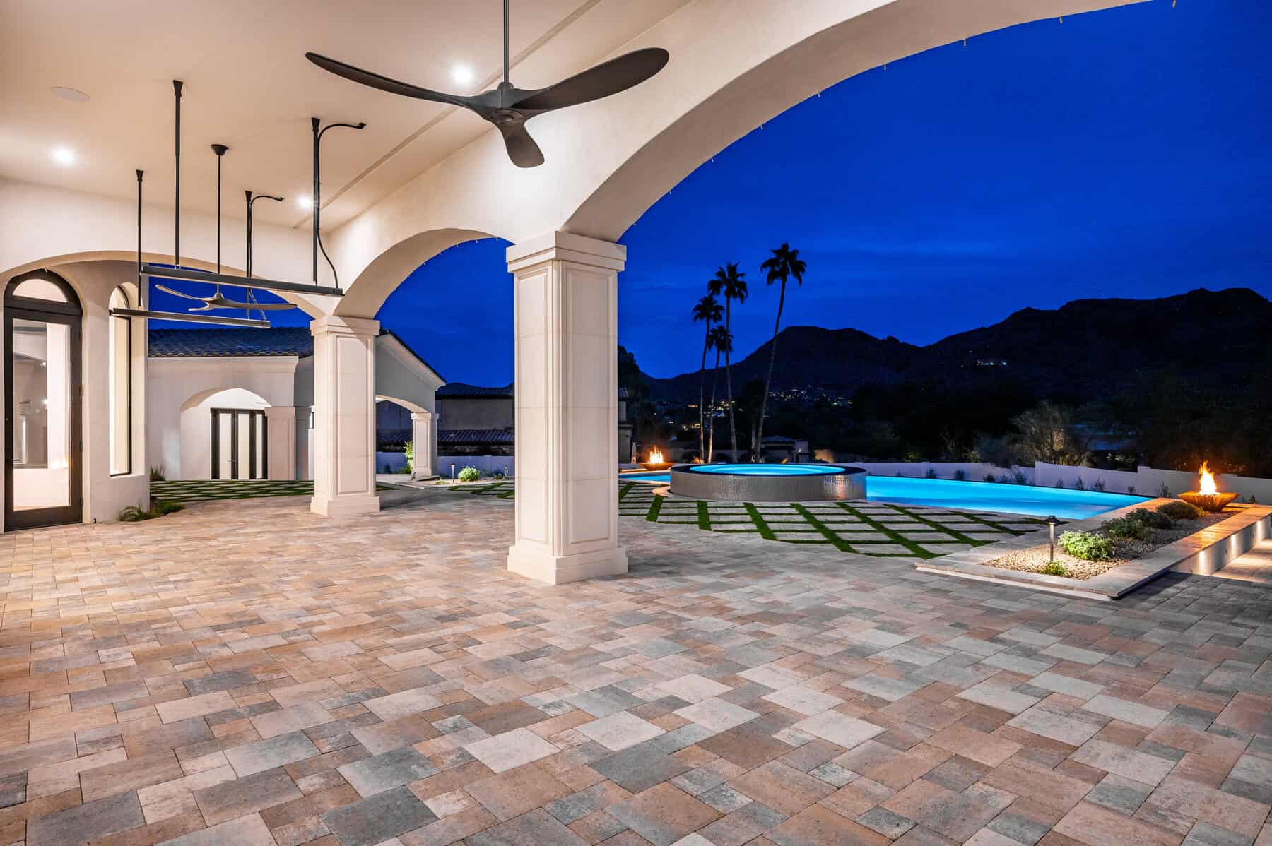 Covered patio with large ceiling fans, arched columns, and stone tile flooring by Avondale Turf and Landscape, overlooking a pool, spa, fire features, and mountain views at dusk with palm trees in the background.