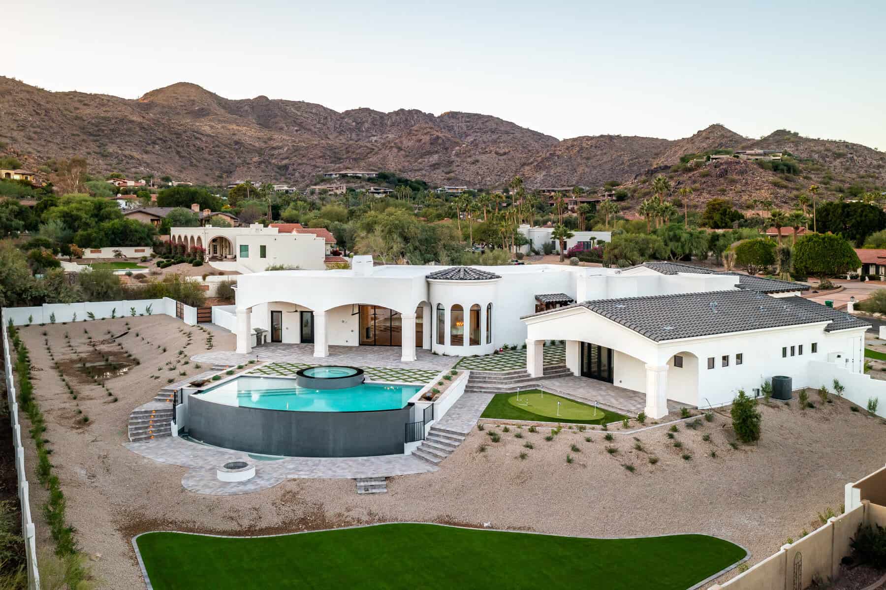 Aerial view of a modern white luxury house with a circular pool, large patio, and landscaped yard by Avondale Turf and Landscape, set against a backdrop of desert hills and neighboring homes.