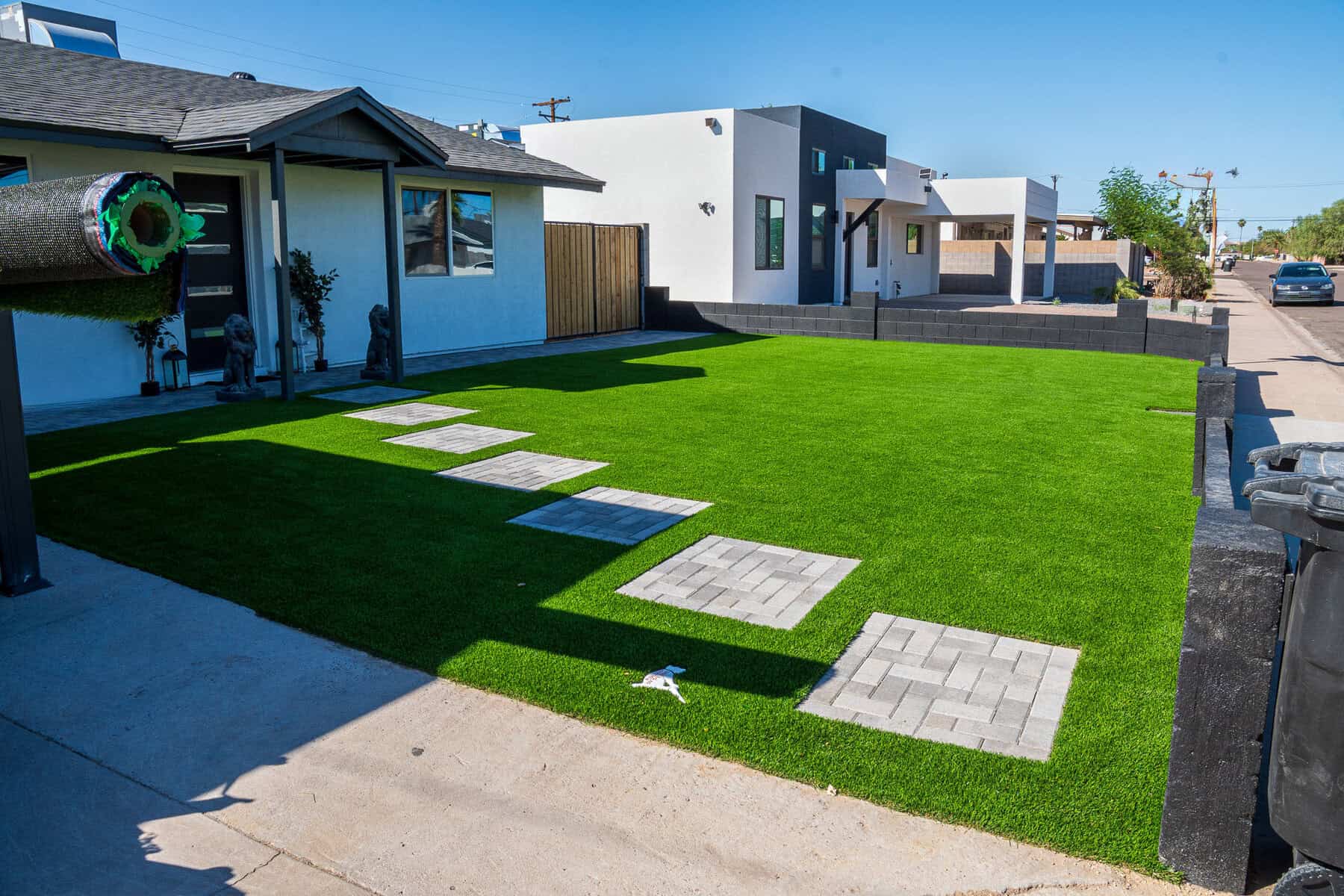 Front yard with bright green artificial turf by Avondale Turf and Landscape, rectangular concrete stepping stones leading to the house, and modern black-and-white homes in a sunny suburban neighborhood. A rolled-up turf section and trash bins are also visible.