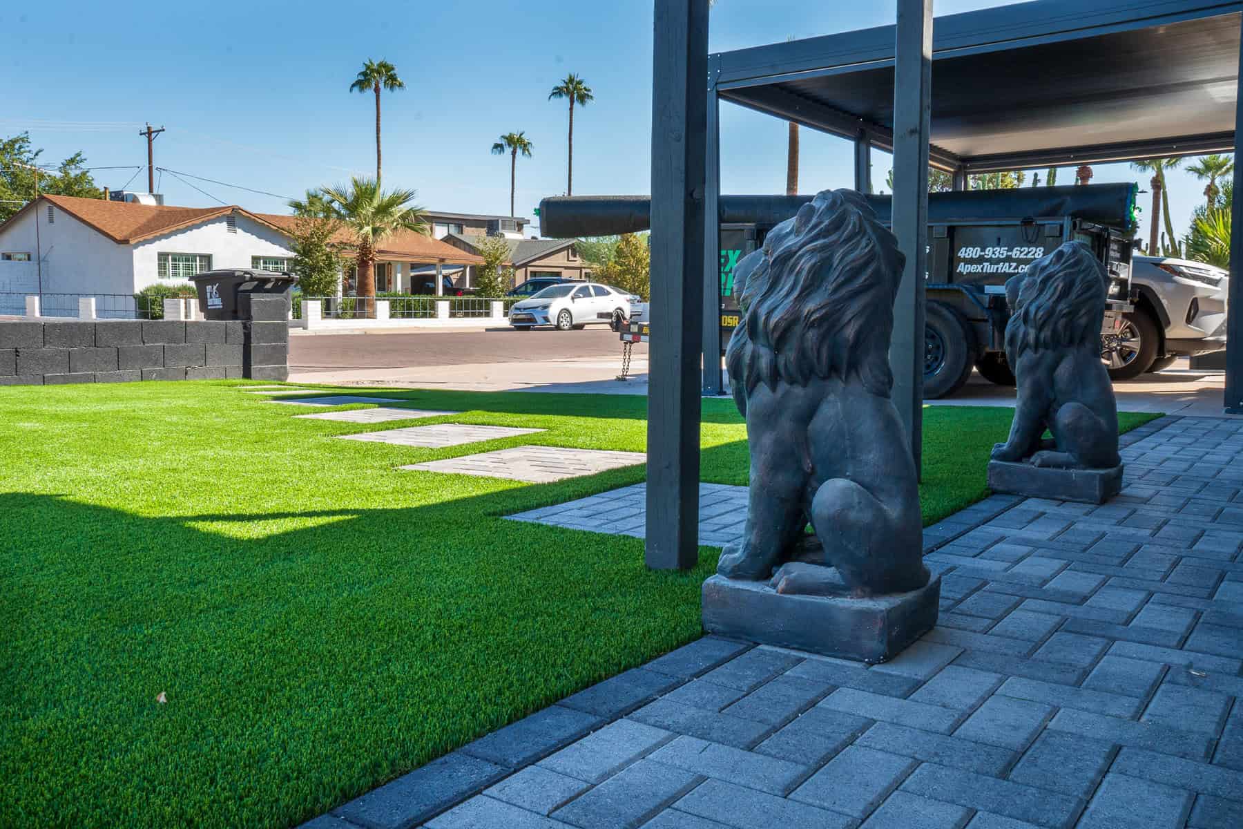 A yard with bright green artificial grass by Avondale Turf and Landscape features stone pavers and two black lion statues at the driveway entrance. Several cars and palm trees are visible in the background under a clear blue sky.