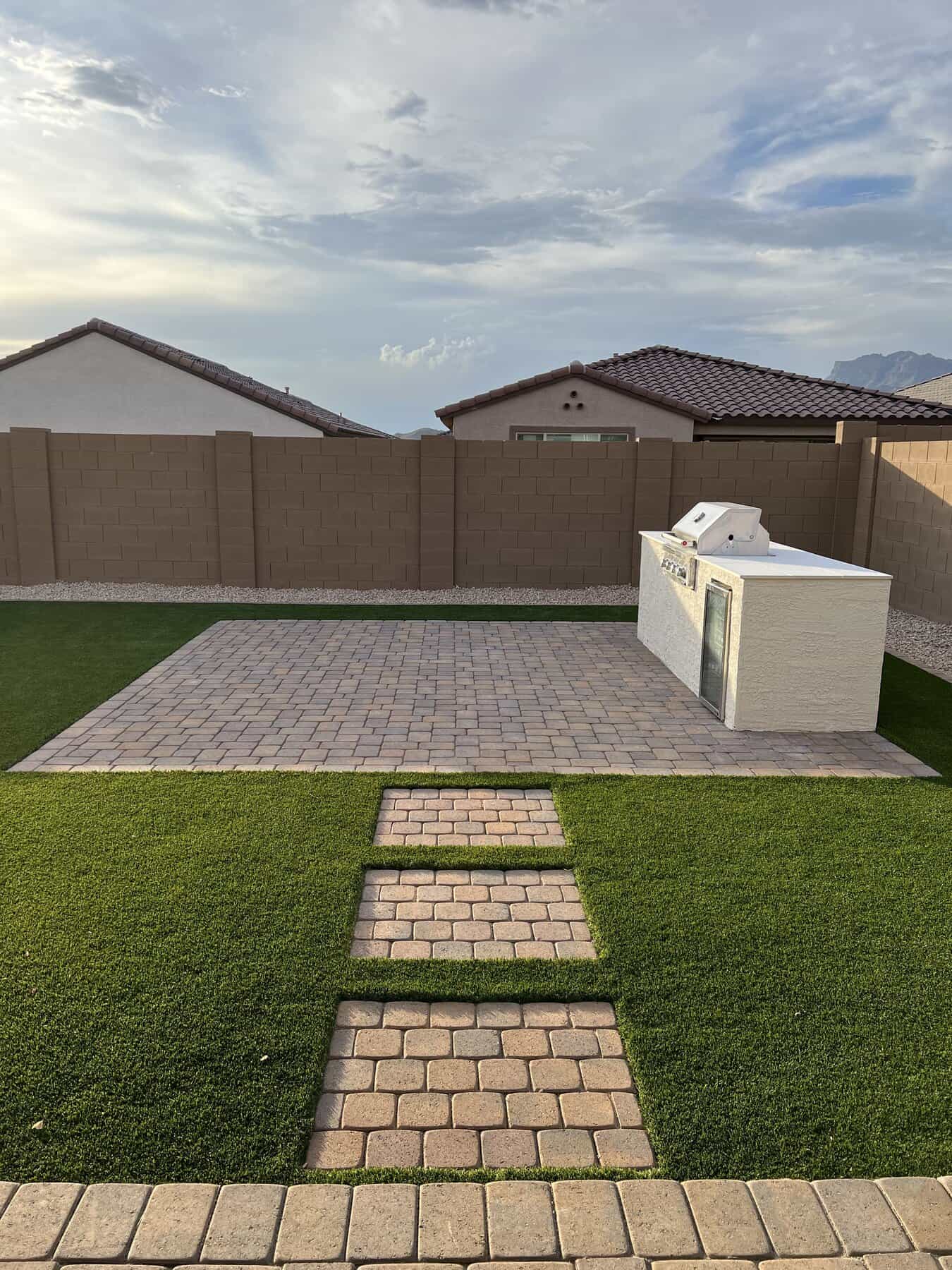 A backyard designed by Avondale Turf and Landscape features a paved patio, built-in outdoor kitchen or grill, lush artificial grass, and stepping stones. The space is enclosed by a brown brick wall with neighboring houses visible.