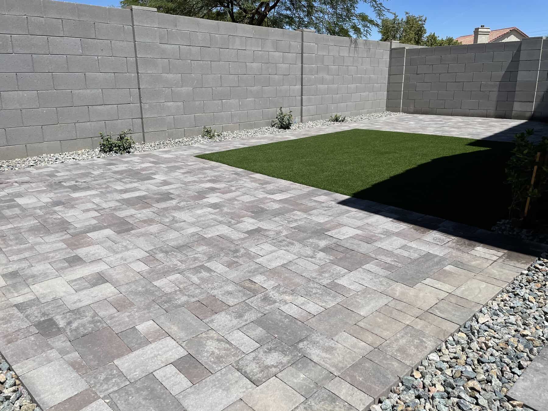 A backyard designed by Avondale Turf and Landscape features gray stone pavers, a small rectangular patch of artificial grass, and a surrounding gravel border, all enclosed by tall gray concrete block walls with a few small plants along the sides.