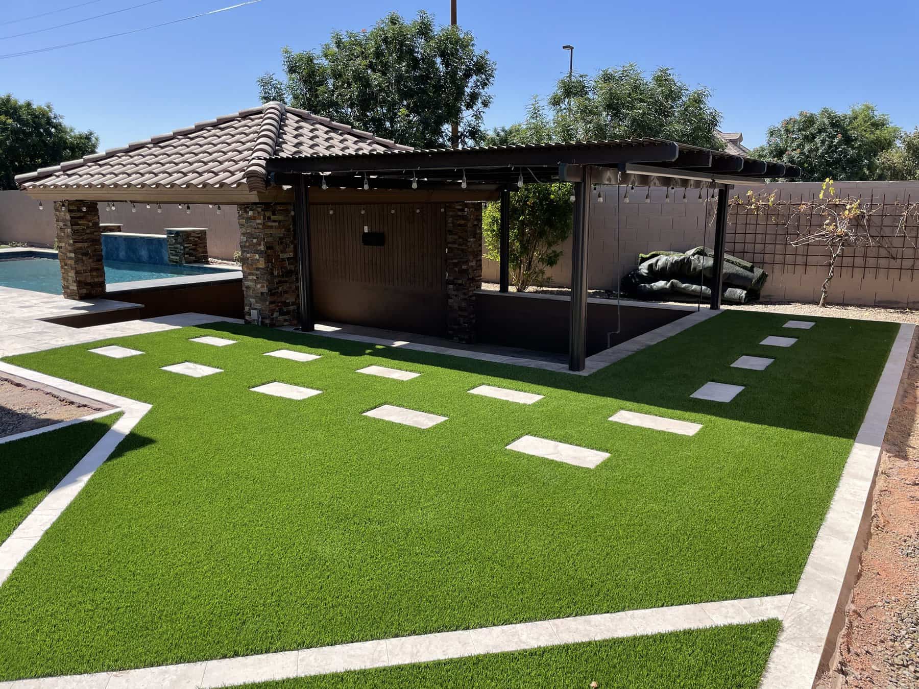 A backyard by Avondale Turf and Landscape with artificial grass, rectangular stone pavers forming paths, a covered patio with a tile roof and stone columns, a pool, and a privacy wall accented by trees and shrubs.