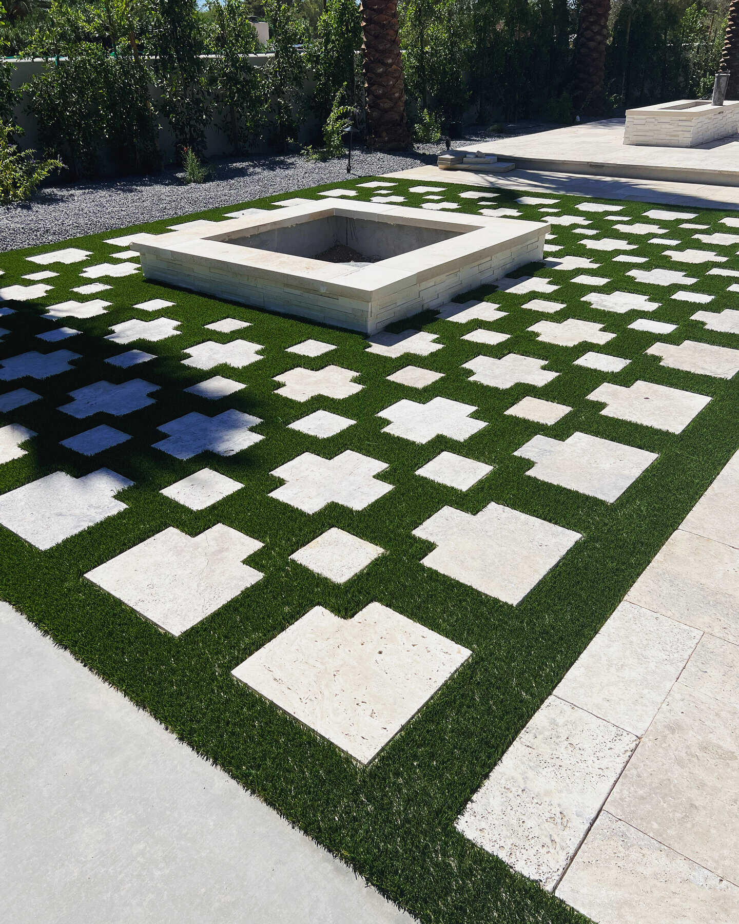 A modern patio by Avondale Turf and Landscape features a square fire pit center, surrounded by artificial grass with square and rectangular stone pavers in a geometric pattern. Trees and shrubs border the area.