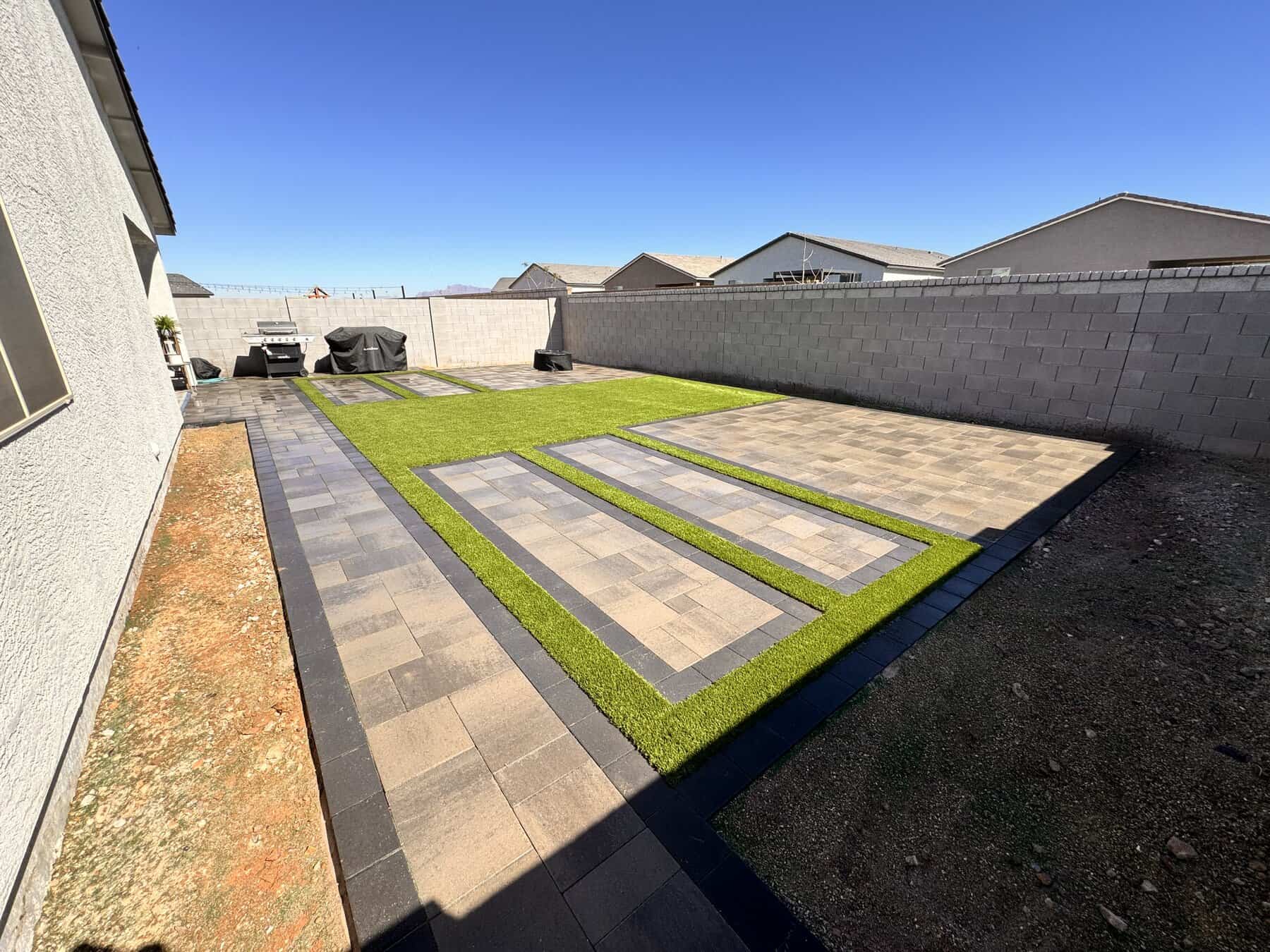 A modern backyard by Avondale Turf and Landscape features rectangular sections of artificial grass and light-colored pavers, bordered by dark bricks, surrounded by a concrete block wall next to a white house under a clear blue sky.