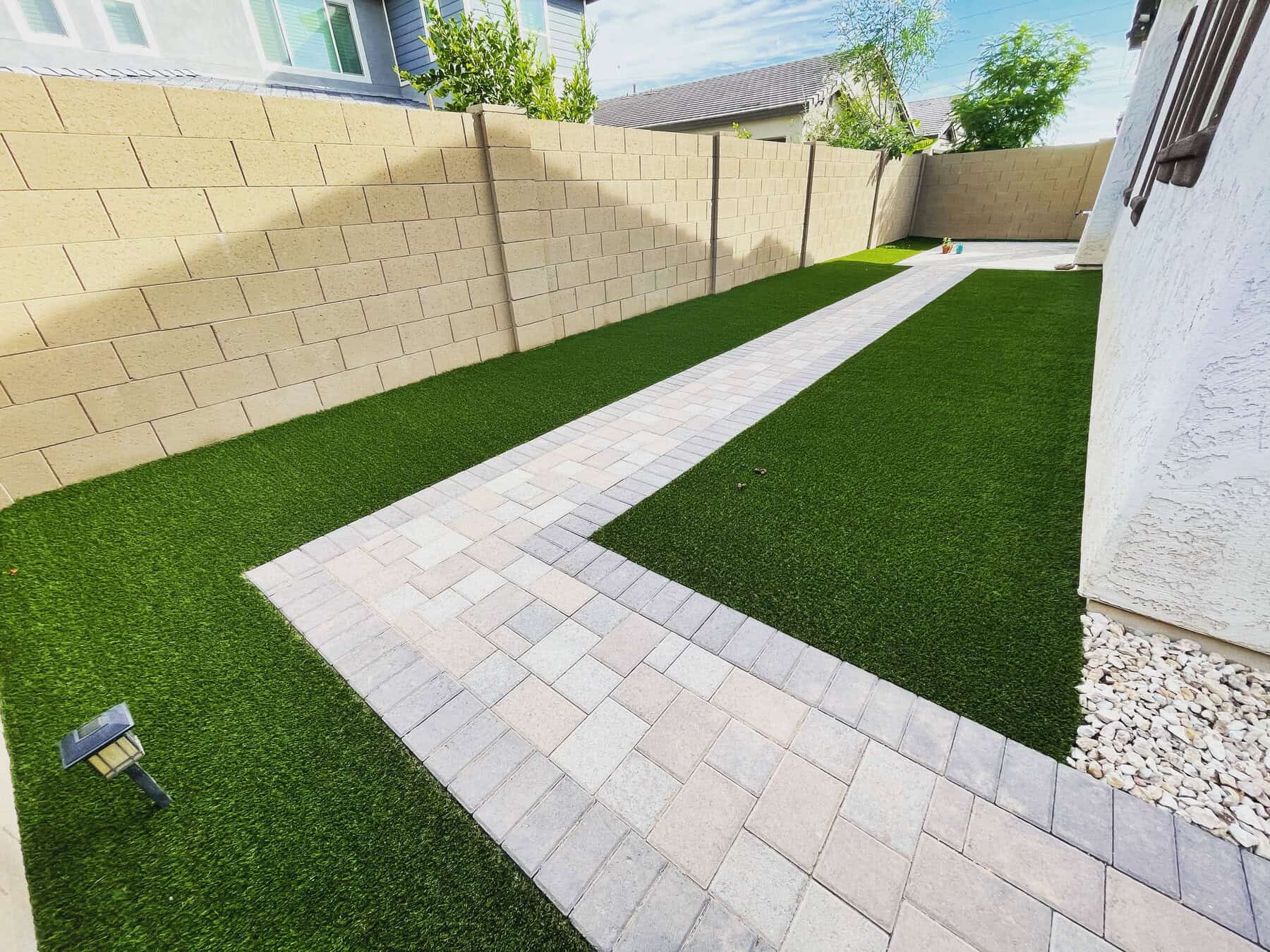 A side yard by Avondale Turf and Landscape features artificial grass, a light-colored stone walkway down the center, tan block walls, and white rocks beside a white house exterior.