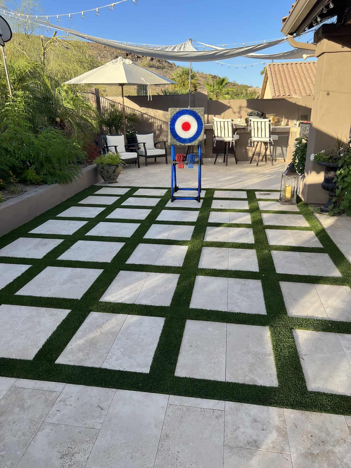 A backyard patio by Avondale Turf and Landscape features a checkered pattern of tiles and artificial grass, outdoor seating, dining area, string lights, and an archery target in the center beneath sun shades.