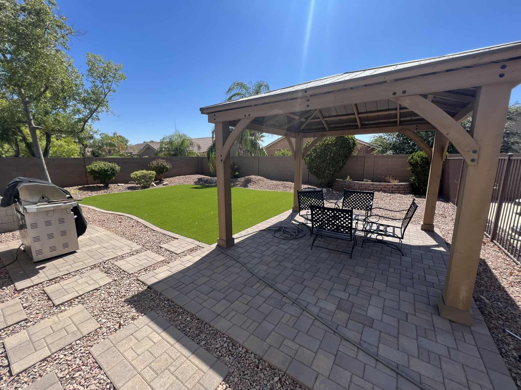 A backyard designed by Avondale Turf and Landscape features a wooden gazebo over a patio table and chairs, a grill to the left, artificial grass in the center, stone pathways, and lush trees and shrubs along a brick wall under a clear blue sky.