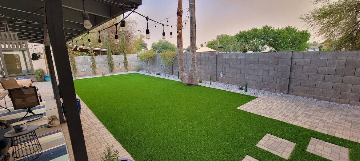 A backyard designed by Avondale Turf and Landscape features artificial grass, brick pavers, string lights, palm trees, and a stone wall fence. Patio furniture sits on a covered patio to the left, while trees and plants line the edge of the yard.