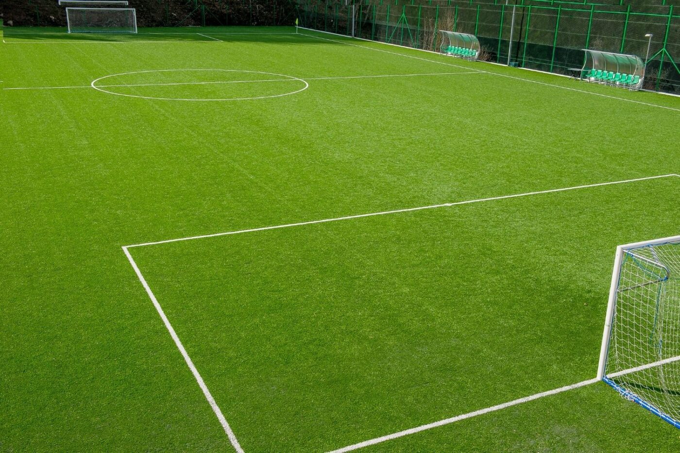 A view of an empty soccer field with green artificial turf by Avondale Turf and Landscape, featuring white boundary lines, two goals, and team benches along the sideline behind a green fence.