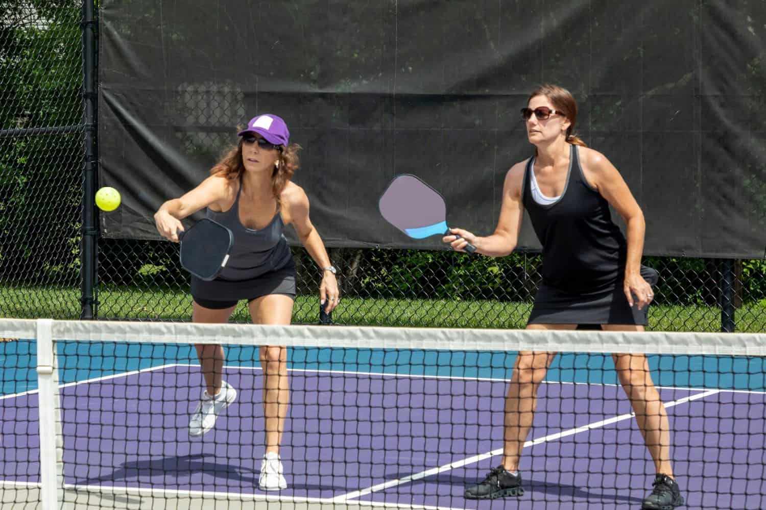 Two women play pickleball on an outdoor court featuring Avondale Turf and Landscape. One, in a purple cap and black tank top, reaches to hit the ball while the other, in sunglasses and a black dress, stands ready with her paddle.