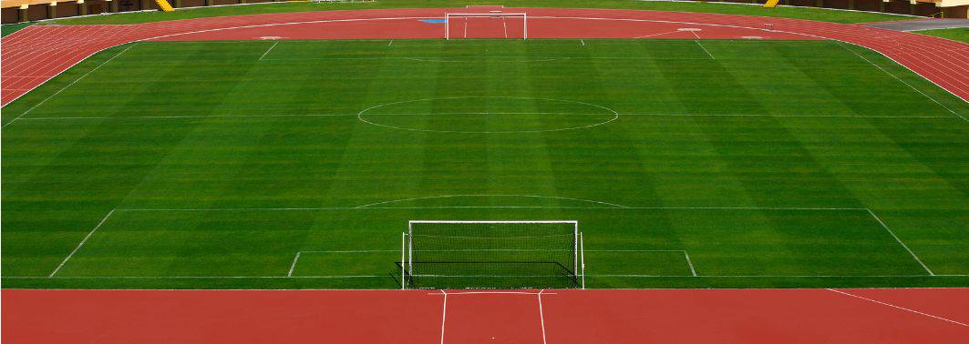 An empty outdoor football (soccer) field with bright green Avondale Turf and Landscape grass, surrounded by a red running track and goals at both ends, viewed from the stands behind one goal.