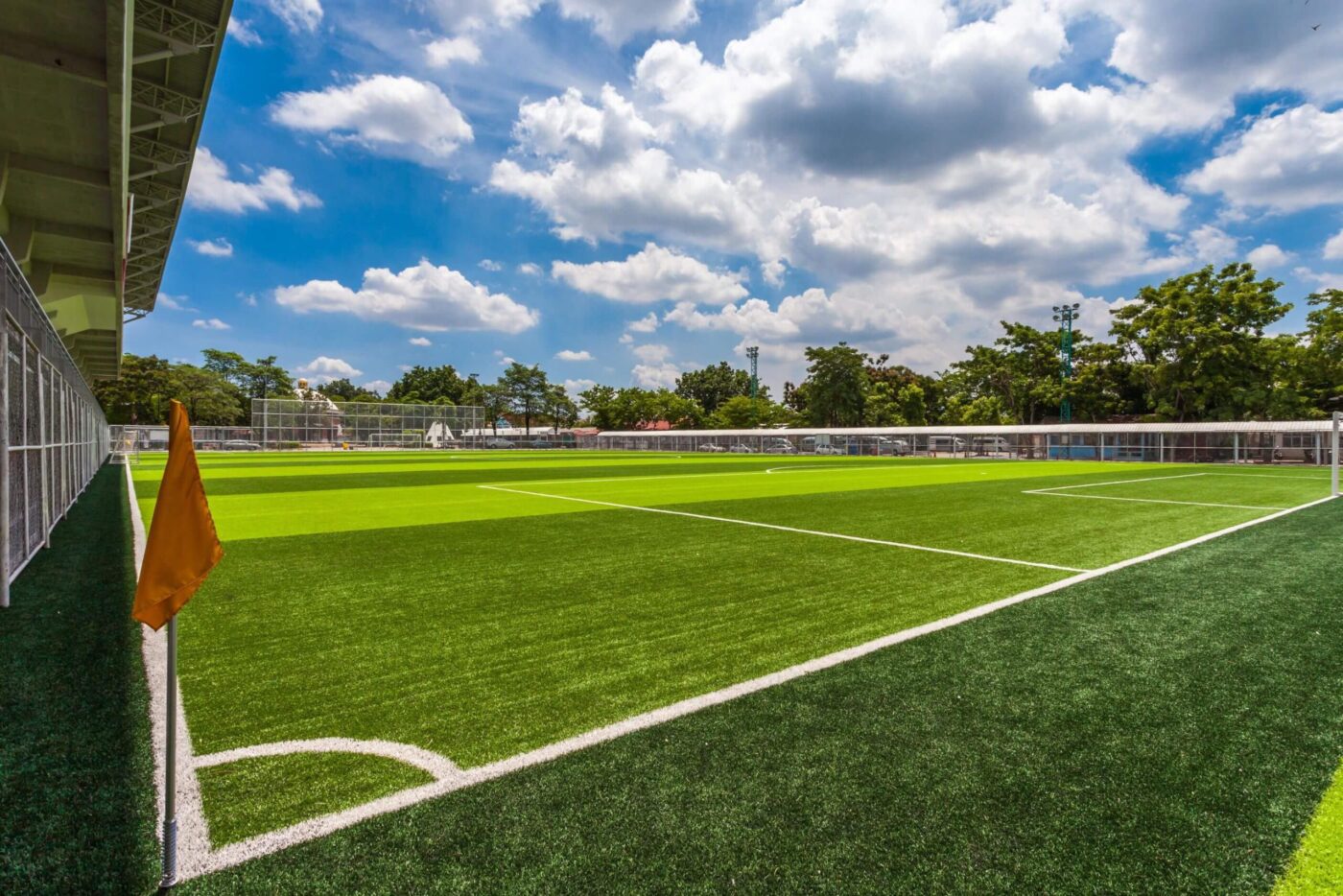 A bright, sunny day at an empty outdoor soccer field with green artificial turf by Avondale Turf and Landscape, white markings, a corner flag, and trees and buildings in the background under a partly cloudy sky.
