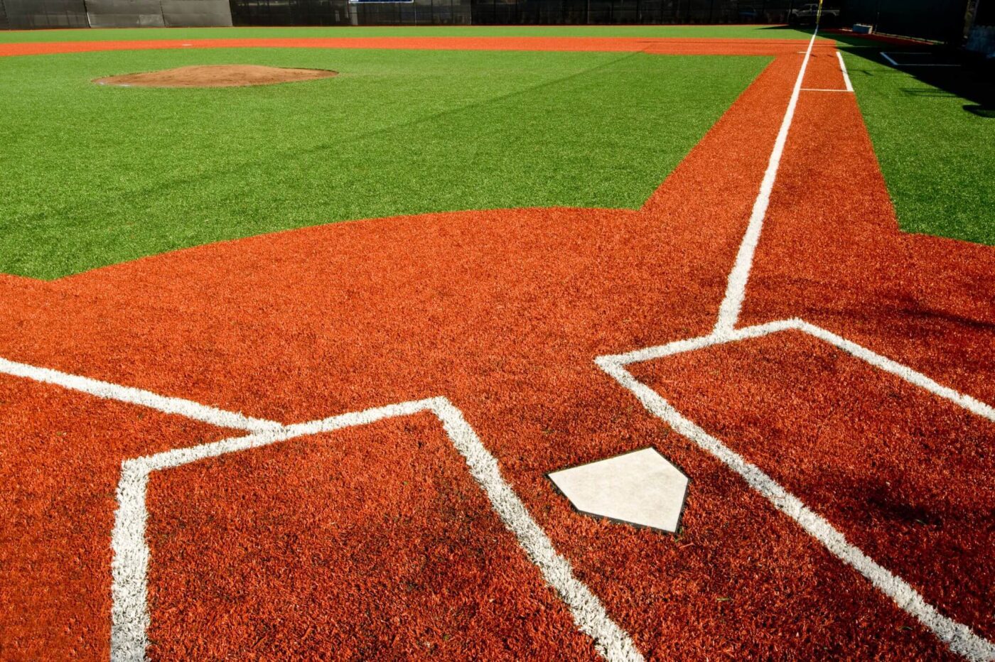 A close-up view of a baseball field's home plate area with bright red artificial turf by Avondale Turf and Landscape, crisp white chalk lines, and the pitcher’s mound visible in the distance.