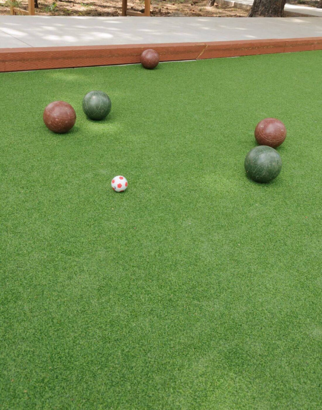 Four bocce balls—two green and two brown—and a small white and red pallino rest on a green Avondale Turf and Landscape artificial turf court, framed by a wood border and trees in the background.