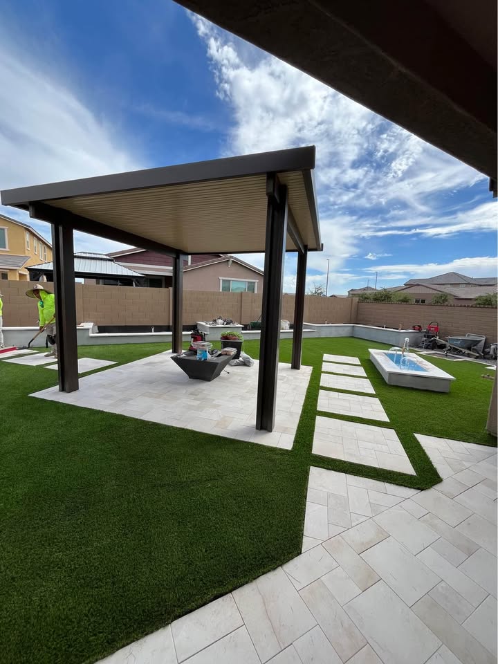 A backyard with artificial grass features a modern pergola on a tiled patio, Pavers Installation Avondale touches, a concrete fire pit, outdoor furniture, and a small fountain. Houses and a blue sky with clouds are visible in the background.