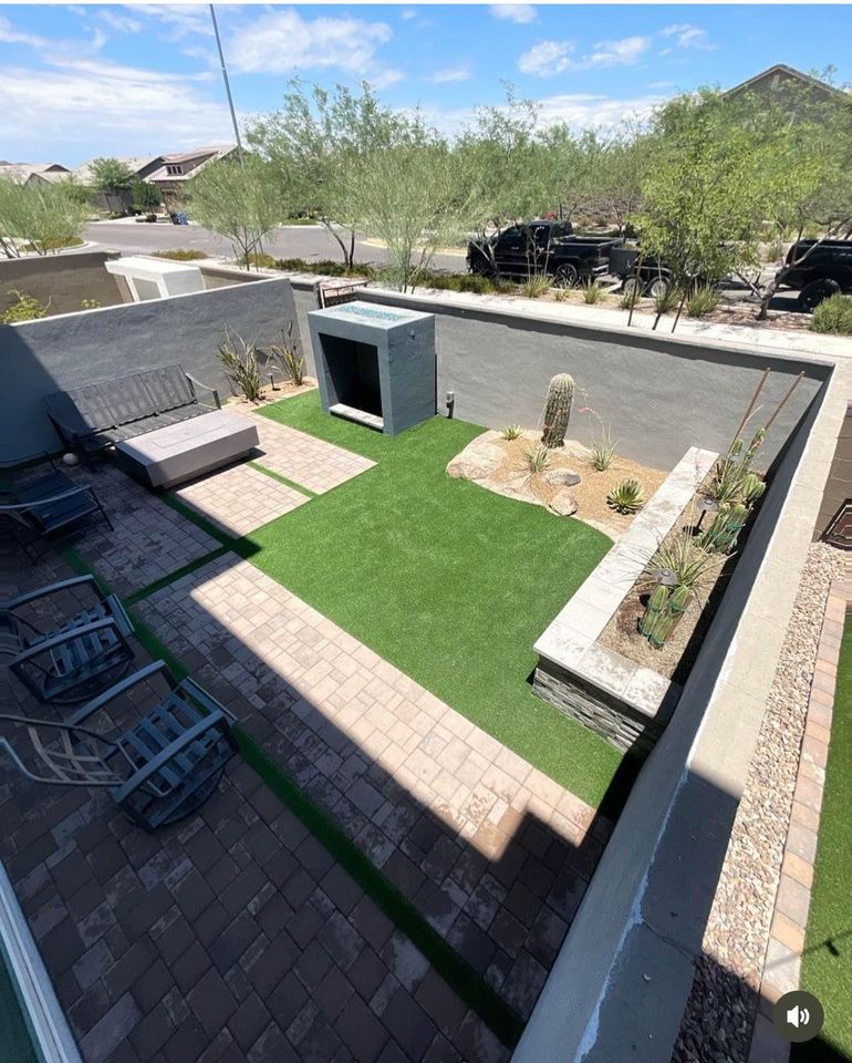 A modern backyard by an expert Artificial Grass Installer Goodyear AZ, featuring artificial grass, a paver patio, outdoor fireplace, seating areas, desert plants, and a stone planter, all enclosed by sleek gray walls on a sunny day.