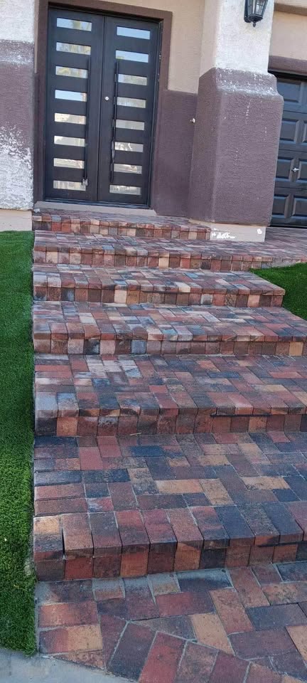 A set of brick steps with an uneven, warped design leads up to a modern dark door with horizontal glass panels. Expertly installed by an Artificial Grass Installer in Glendale AZ, lush artificial grass borders the steps on the left side.