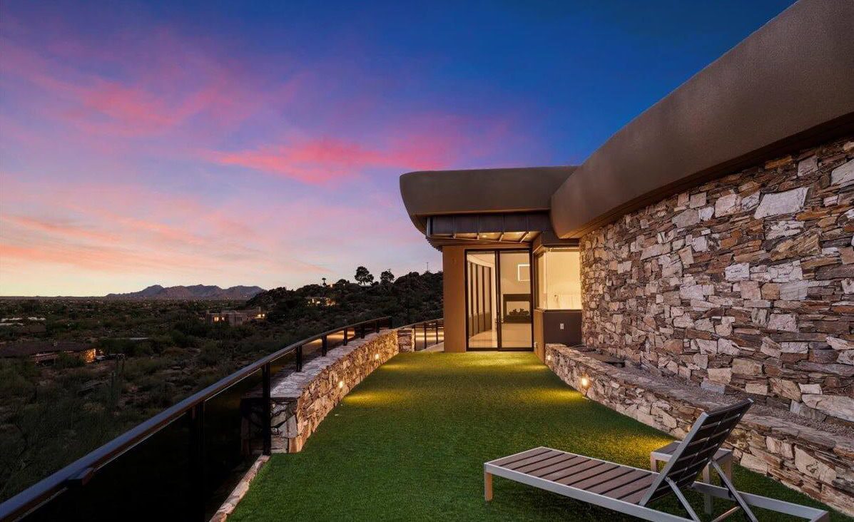 A modern building with a stone exterior and curved balcony overlooks a scenic landscape at sunset. Installed by an expert Artificial Grass Installer Laveen AZ, the balcony features lush artificial grass flooring, ground lighting, and distant mountains.