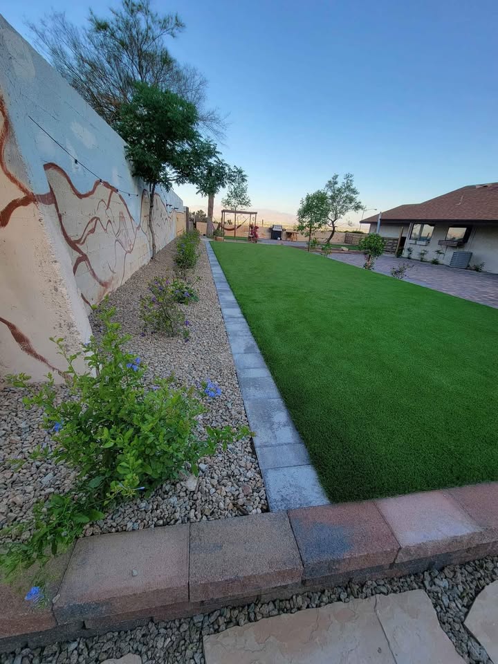 A landscaped backyard with green artificial turf installed by a trusted Artificial Grass Installer Glendale AZ, a stone border, gravel beds with small plants, a mural on the left wall, and a patio area near a house under a clear blue sky.