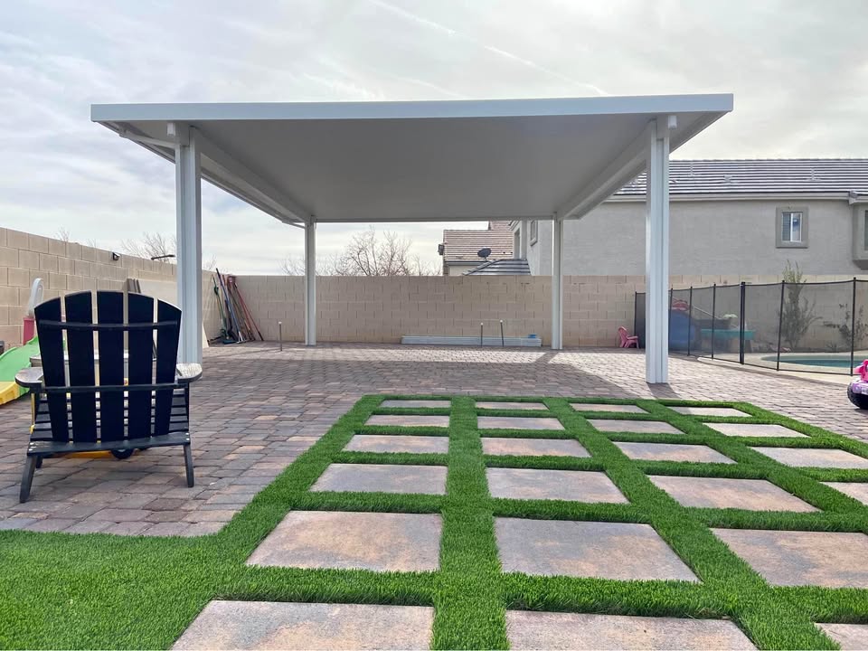 A backyard with a modern white patio cover, paved stone flooring, artificial grass squares by an expert Artificial Grass Installer Laveen AZ, a black Adirondack chair, and a fenced pool area against a tall beige brick wall and neighboring houses.